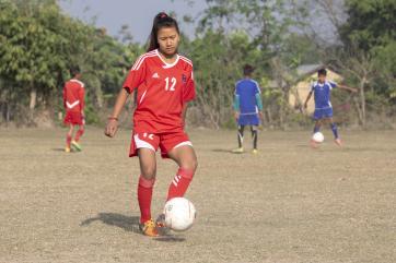 A young person in a red uniform kicking a ball on a practice field.