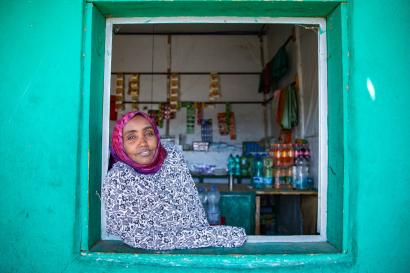 A shopkeeper and pastoralist in a window with goods and supplies behind them.