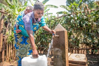 Mother, with child in sling on her back, collects water at outdoor water spigot.