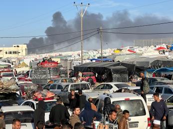 People standing in between cars and tents as smoke bellows in the background