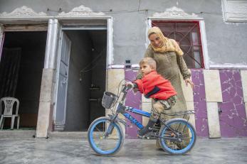 A boy in syria rides a bicycle as a women observes.
